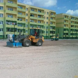 Żółta ładowarka Volvo z niebieskim osprzętem równającym teren z jasnego kruszywa na tle wielopiętrowych bloków mieszkalnych; w tle widoczny walec drogowy.