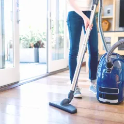 Osoba odkurza drewnianą podłogę w jasnym wnętrzu, widoczny niebieski odkurzacz cylindryczny, drzwi balkonowe i fragment mebli.