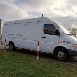 Biały bus transportowy marki Mercedes Sprinter zaparkowany na poboczu, z widocznym numerem telefonu na boku, na tle zachmurzonego nieba i drzew bez liści.