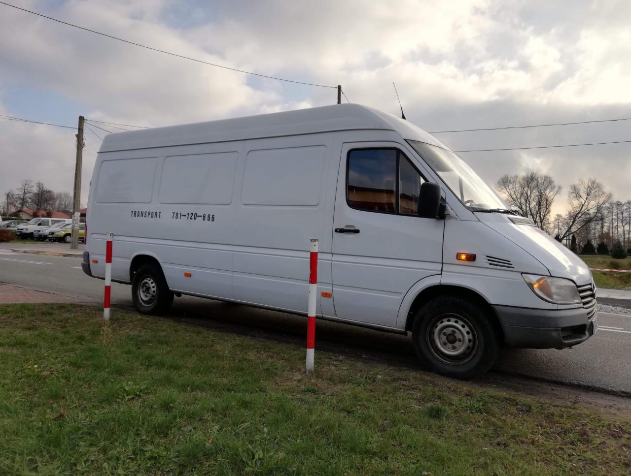 Biały bus transportowy marki Mercedes Sprinter zaparkowany na poboczu, z widocznym numerem telefonu na boku, na tle zachmurzonego nieba i drzew bez liści.
