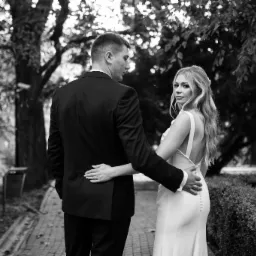 Czarno-białe zdjęcie pary młodej, mężczyzna w garniturze obejmuje kobietę w sukni ślubnej z odkrytymi plecami, oboje stoją tyłem na tle drzew i krzew&oacute;w w parku, kobieta spogląda przez ramię.