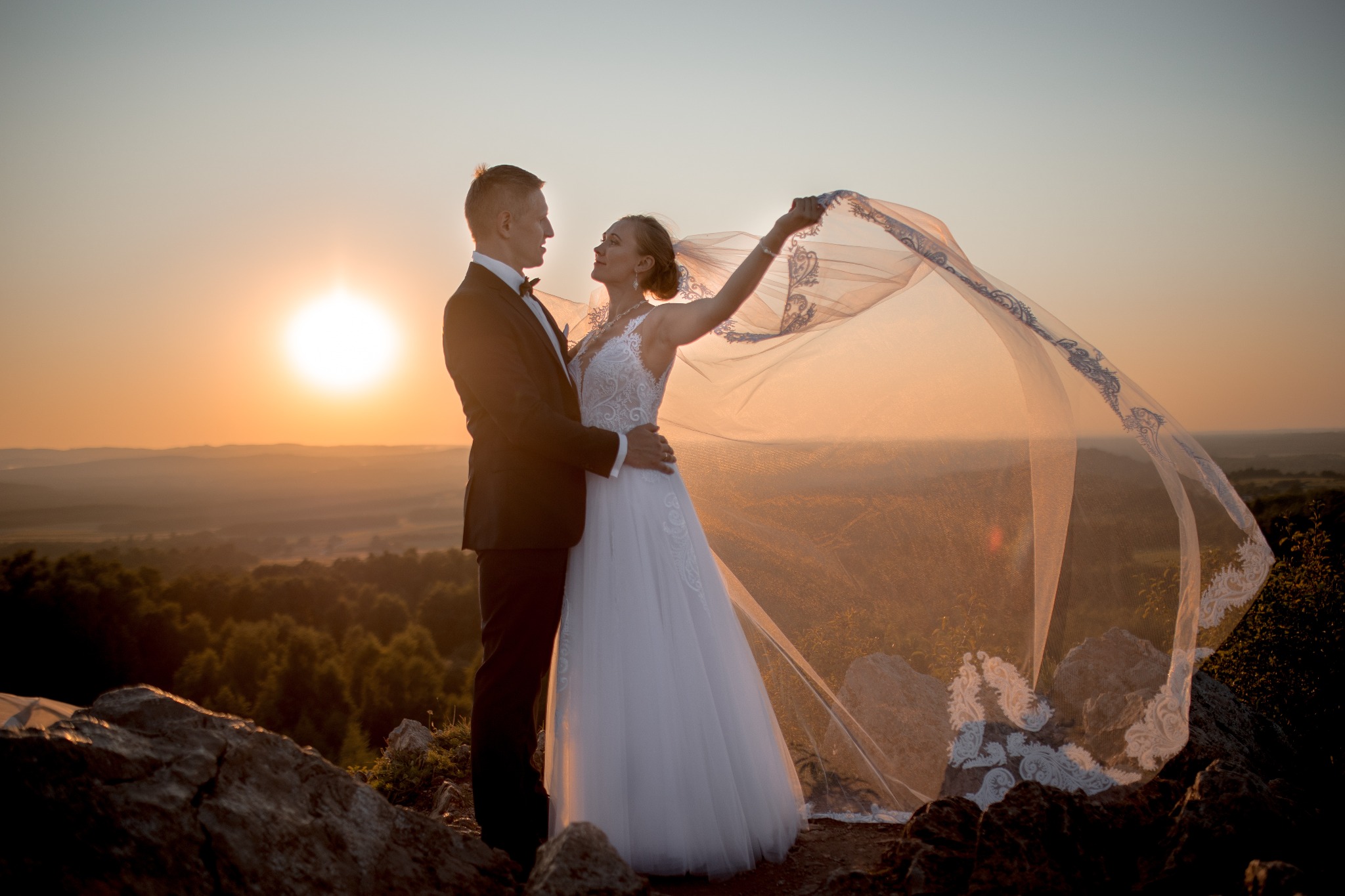 Para młoda na tle zachodzącego słońca, panna młoda trzyma welon z niebieskim haftem, stoją na skalistym wzgórzu.