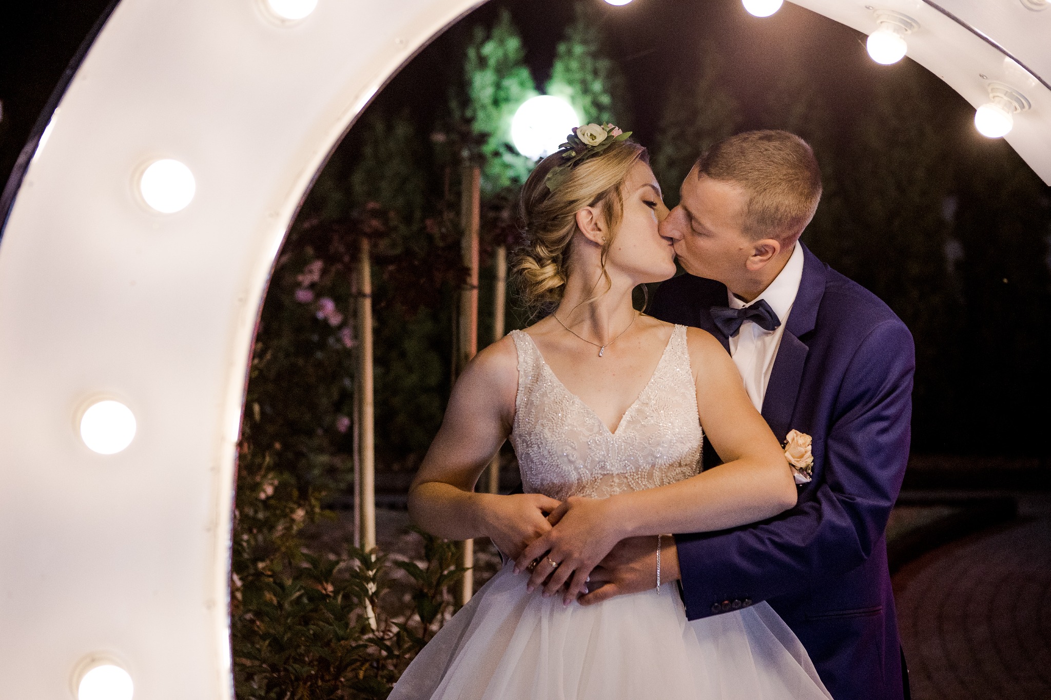 Pocałunek nowożeńców pod oświetloną bramą w nocy, panna młoda w sukni z aplikacjami i wiankiem, pan młody w granatowym smokingu z butonierką.