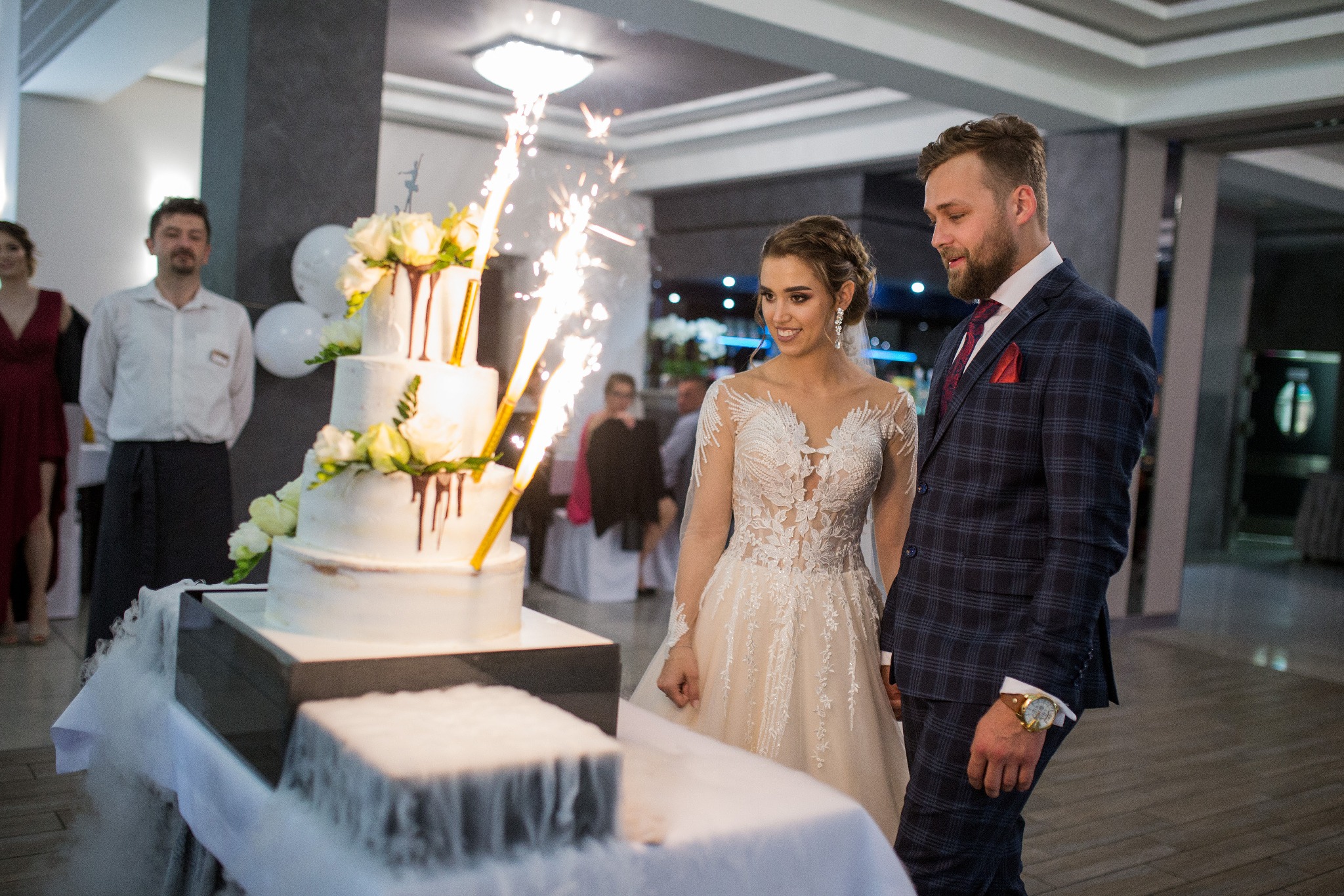 Para młoda stoi przed tortem weselnym z fontannami iskier i dekoracjami z białych róż, widoczny dym z suchego lodu oraz goście w tle.