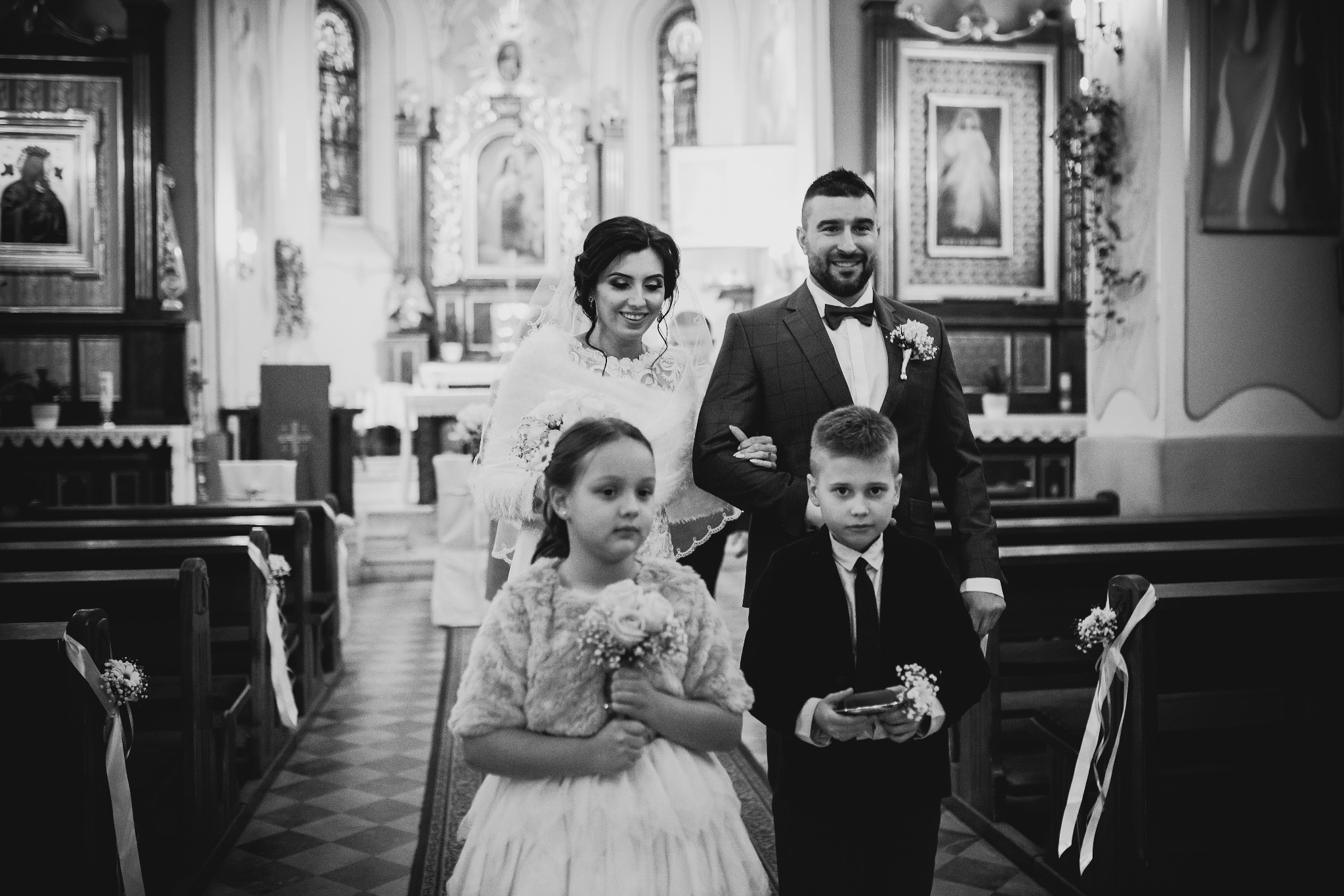Czarno-białe zdjęcie pary młodej idącej w kościele, z dwójką dzieci niosących kwiaty, dekoracje kwiatowe na ławkach.