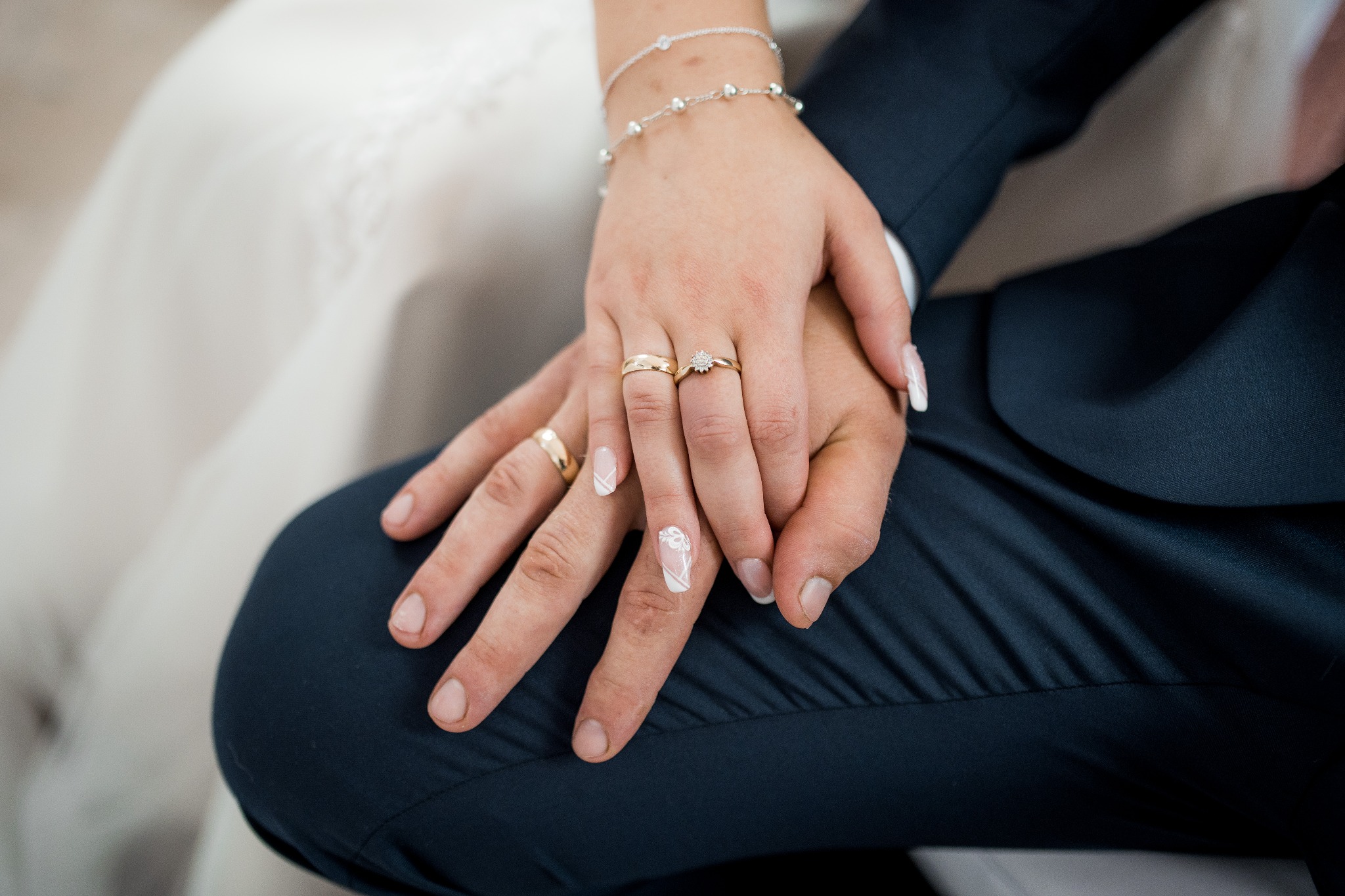 Dłonie pary młodej splecione na tle granatowego garnituru, widoczne złote obrączki i delikatna bransoletka.