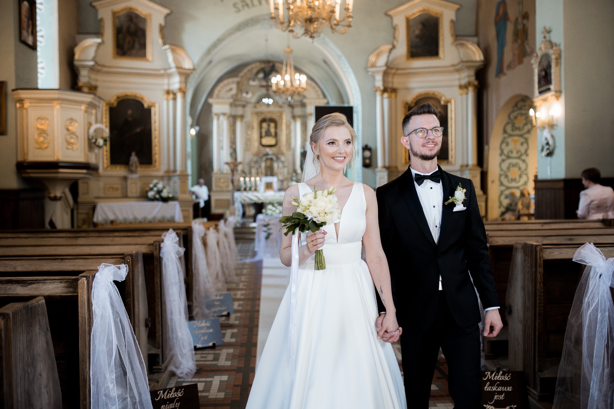 Para młoda trzymająca się za ręce w kościele, panna młoda w białej sukni z bukietem białych róż, pan młody w czarnym smokingu z muszką, w tle drewniane ławki z dekoracjami z tiulu.