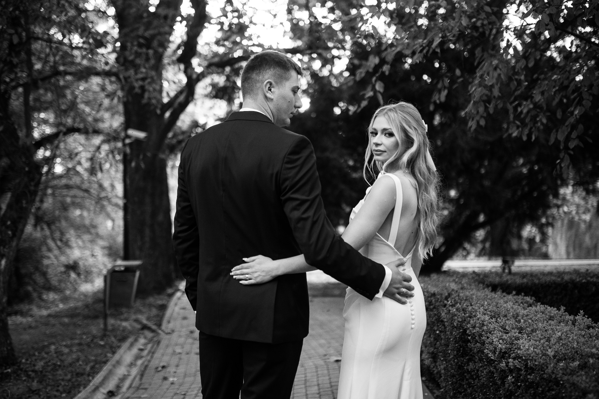 Czarno-białe zdjęcie pary młodej, mężczyzna w garniturze obejmuje kobietę w sukni ślubnej z odkrytymi plecami, oboje stoją tyłem na tle drzew i krzewów w parku, kobieta spogląda przez ramię.