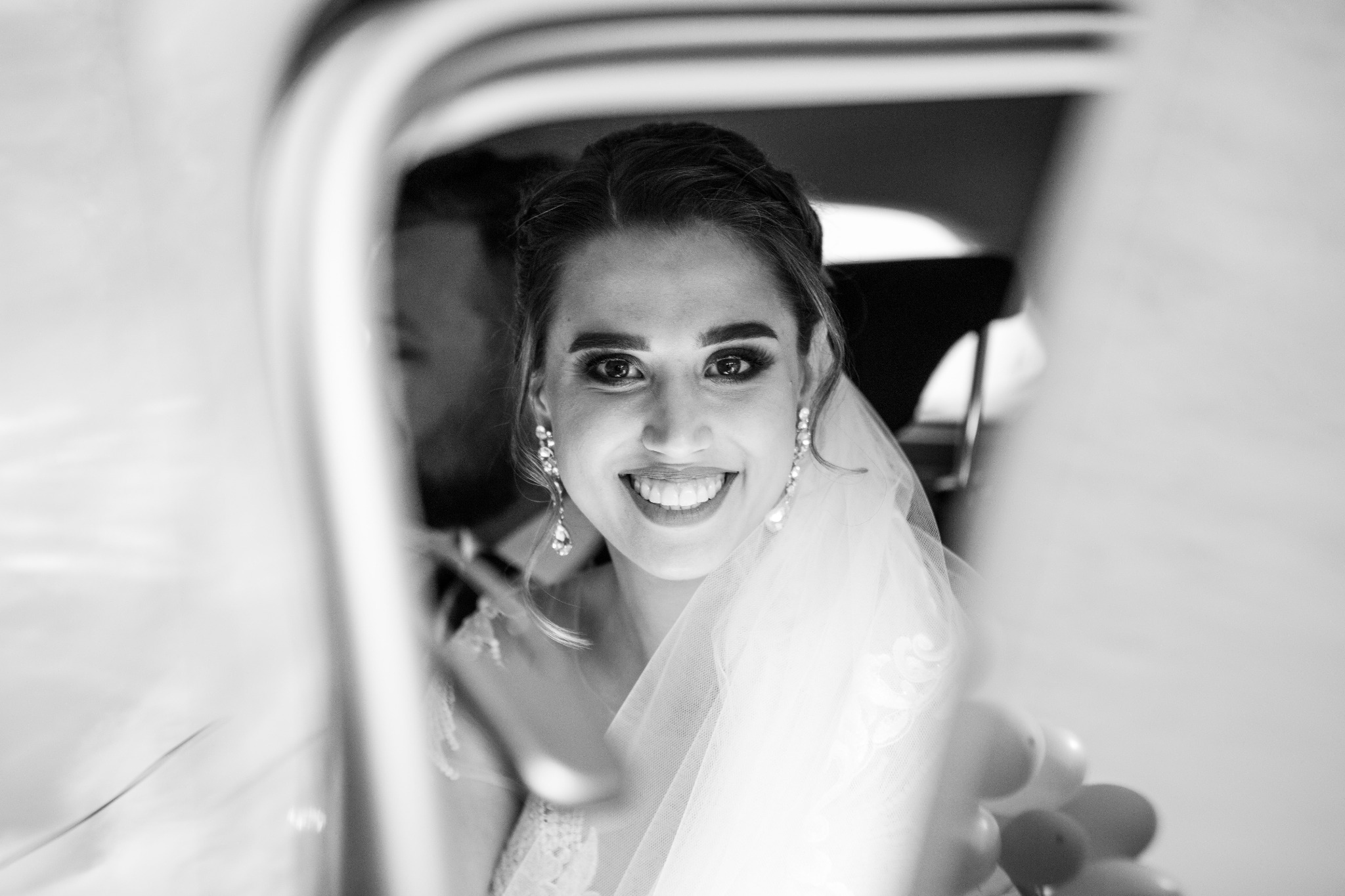 Czarno-białe zbliżenie uśmiechniętej panny młodej w welonie i z wiszącymi kolczykami, widziana przez otwarte drzwi samochodu, z widocznymi balonami w tle.
