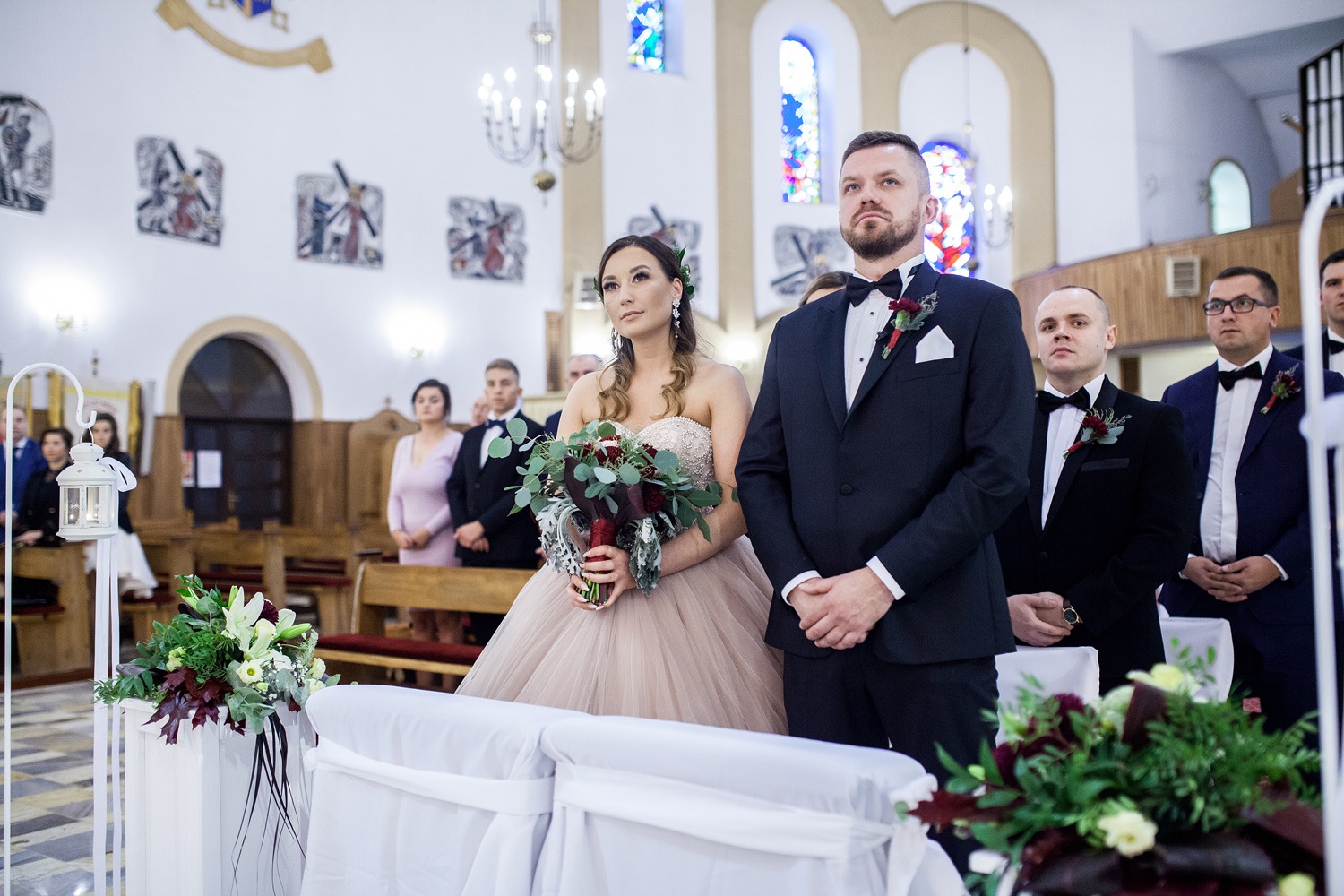 Para młoda w kościele podczas ceremonii ślubnej, panna młoda trzyma bukiet z ciemnymi akcentami, w tle goście ubrani w garnitury i sukienki.
