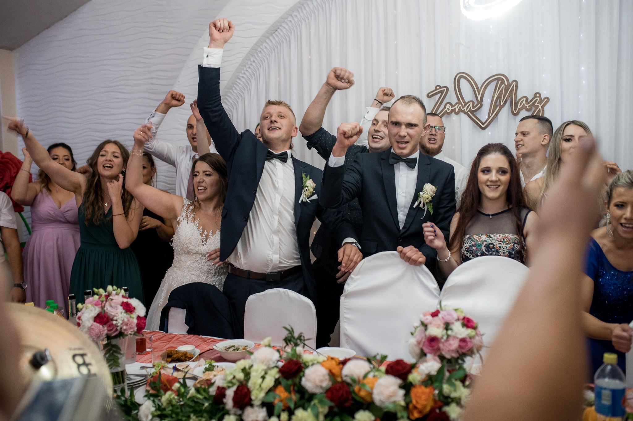 Rozradowani goście weselni, w tym para młoda w sukni z koronkową górą i dwóch panów młodych w smokingach z muchami, wznoszą ręce podczas uroczystości, widoczny stół z kwiatami i jedzeniem, w tle...