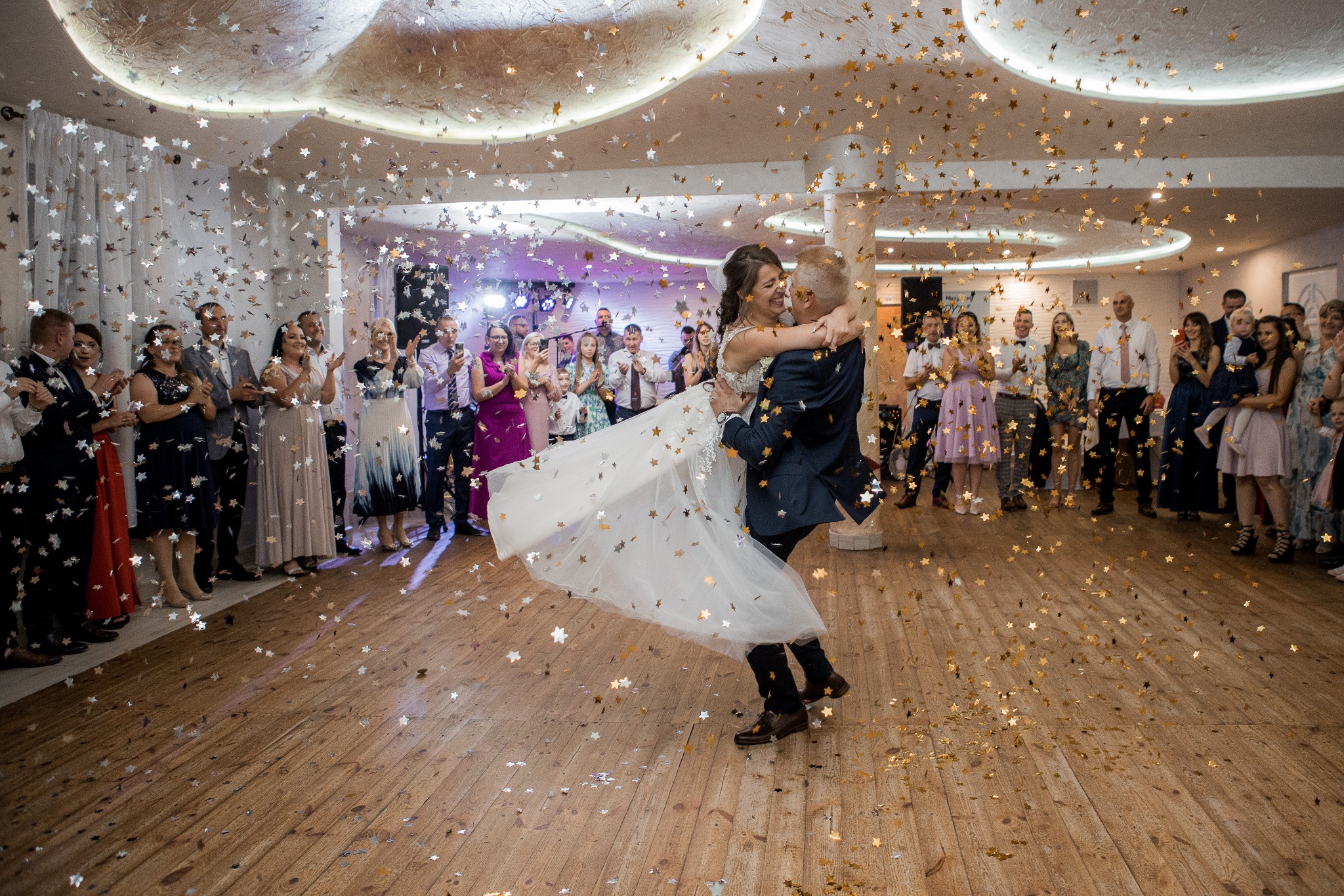 Pierwszy taniec pary młodej w otoczeniu gości i konfetti w kształcie gwiazd na drewnianym parkiecie.