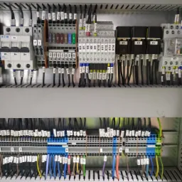 Rozdzielnica elektryczna z widocznymi bezpiecznikami, przekaźnikami i licznymi przewodami oznaczonymi etykietami, uporządkowanymi w listwy zaciskowe.