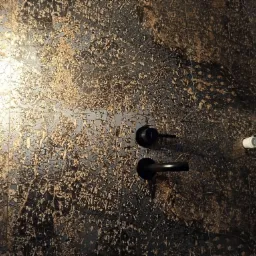 Ściana wyłożona czarno-złotymi płytkami ceramicznymi z wyraźną fakturą, widoczna czarna bateria umywalkowa i biały uchwyt na papier toaletowy.