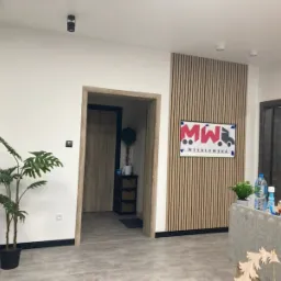 Wnętrze biura z drewnianymi lamelami na ścianie za logo firmy transportowej MW Wesołowska, roślina doniczkowa w lewym dolnym rogu i widok na kolejne pomieszczenie w tle.