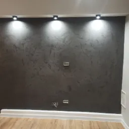 Ściana w salonie pokryta dekoracyjnym, ciemnoszarym tynkiem strukturalnym, oświetlona trzema punktowymi lampami sufitowymi, z widocznymi gniazdkami elektrycznymi i drewnianą podłogą.