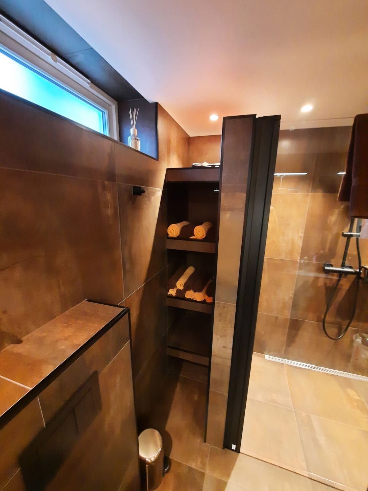 Nowoczesna łazienka z brązowymi płytkami imitującymi rdzę, kabiną prysznicową typu walk-in z czarną ramą i wbudowaną półką z ręcznikami w kolorach ziemi.