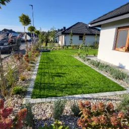 Zadbany trawnik z równo przystrzyżoną trawą, obrzeżony kamiennymi płytami i ozdobnymi rabatami z hortensjami i ozdobnymi trawami, na tle domów jednorodzinnych w słoneczny dzień.