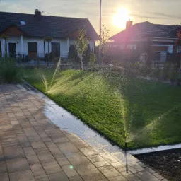 Automatyczny system nawadniania trawnika w ogrodzie, zraszacz w akcji, widok na zielony trawnik, kostka brukowa i dom jednorodzinny w tle, ciepłe światło zachodzącego słońca.