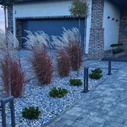 Nowoczesny ogród przydomowy w Witaszycach z ozdobnymi trawami, krzewami iglastymi i kamieniami, oświetlony niskimi lampami wzdłuż ścieżki z kostki brukowej.