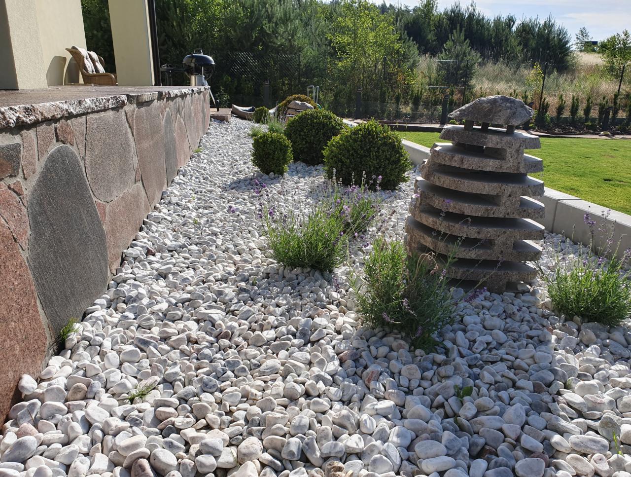 Ogród z białymi kamieniami, ozdobnymi krzewami, lawendą i kamienną rzeźbą w kształcie stosu płyt, widok na trawnik i drzewa w tle.