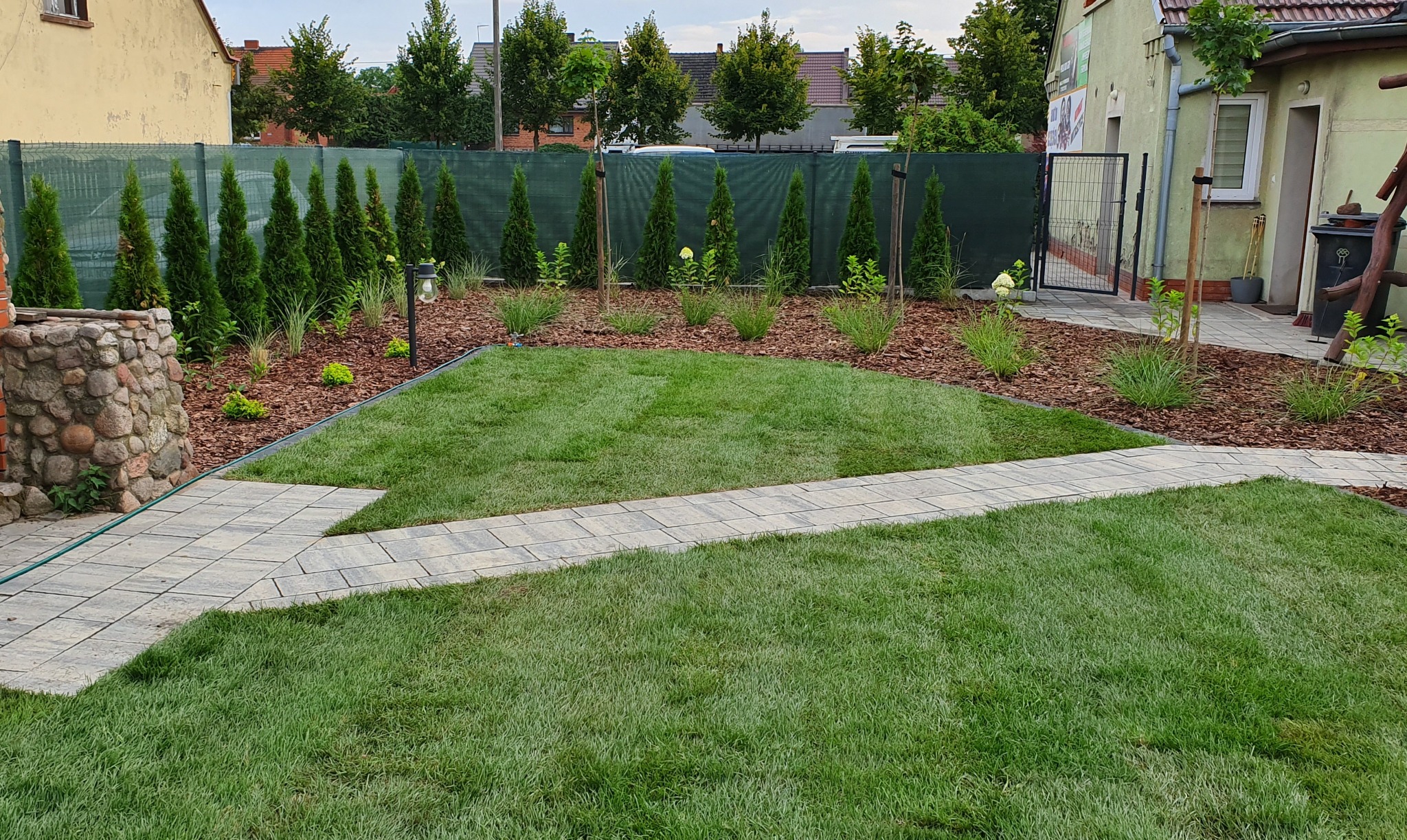 Nowo założony ogród z równo przystrzyżonym trawnikiem, ścieżką z kostki brukowej, obsadzony tujami i krzewami wysypanymi korą, z widocznym kamiennym elementem dekoracyjnym.