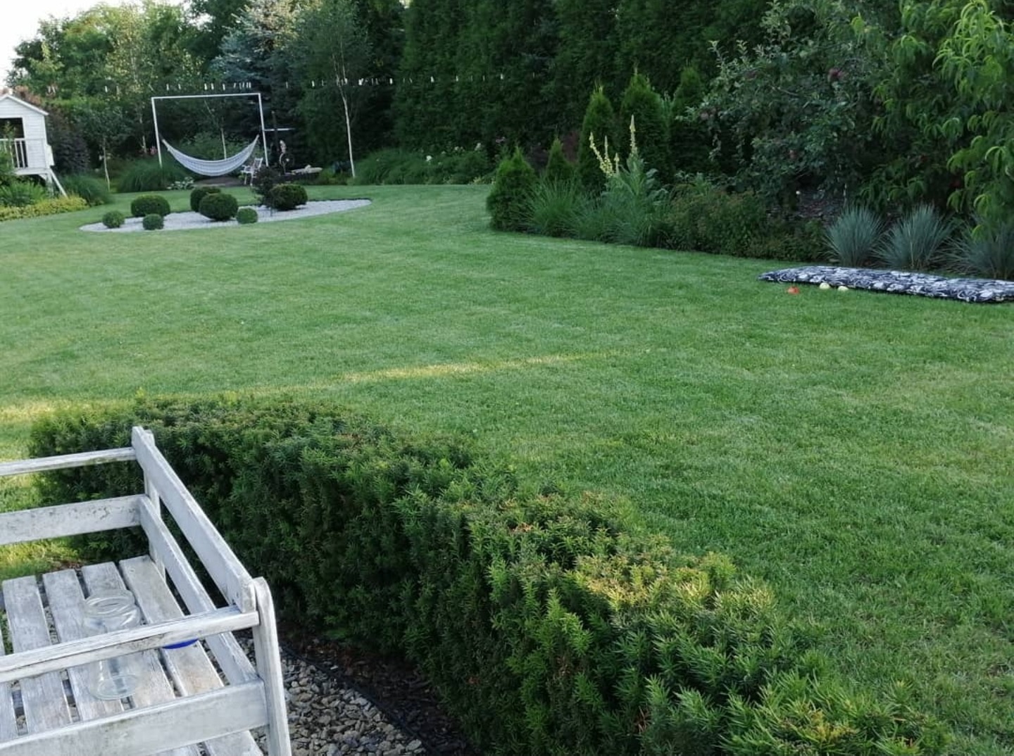 Zadbany trawnik w Witaszycach z równo przyciętym żywopłotem, hamakiem i domkiem dla dzieci w tle, widok z perspektywy ławeczki.
