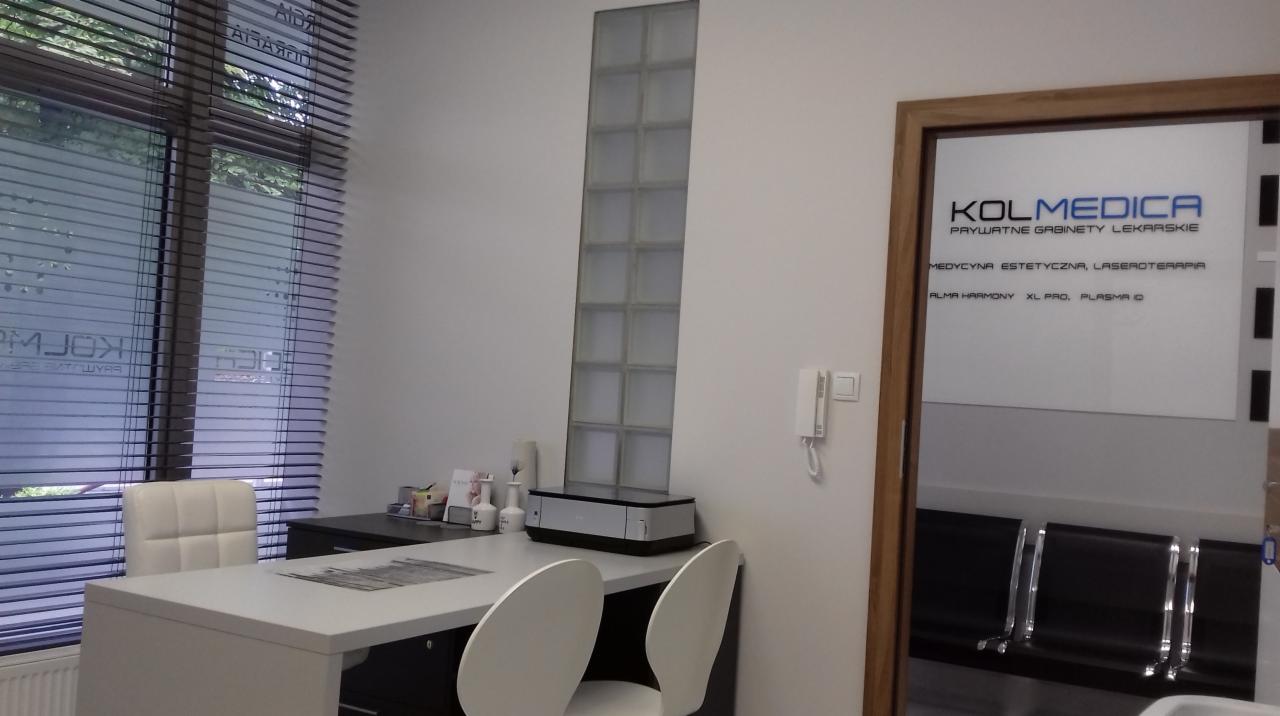 Wnętrze gabinetu lekarskiego z biurkiem, krzesłami, oknem z żaluzjami i widocznym logo firmy KOLMEDICA na szklanych drzwiach.