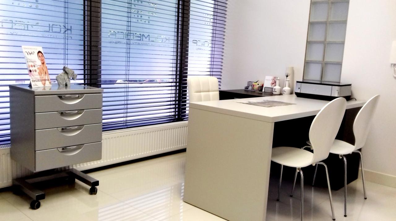 Jasne wnętrze gabinetu z biurkiem, krzesłami, metalową komodą na kółkach z materiałami reklamowymi i oknem z żaluzjami.