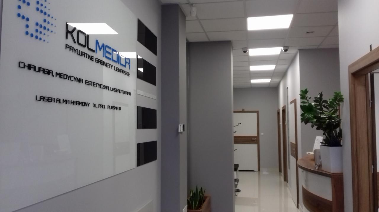 Korytarz w klinice z recepcją, widoczna tablica z nazwą 'KOLMEDICA' i informacjami o usługach: chirurgia, laseroterapia.