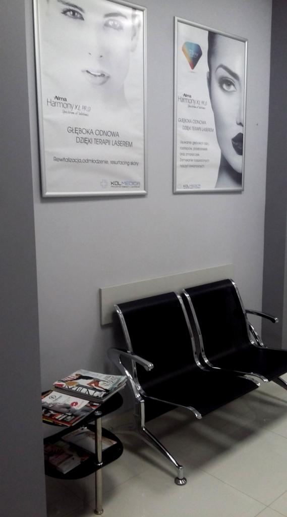 Poczekalnia z dwoma czarnymi krzesłami, stolikiem z czasopismami i plakatami reklamującymi laserowe zabiegi na ścianie.