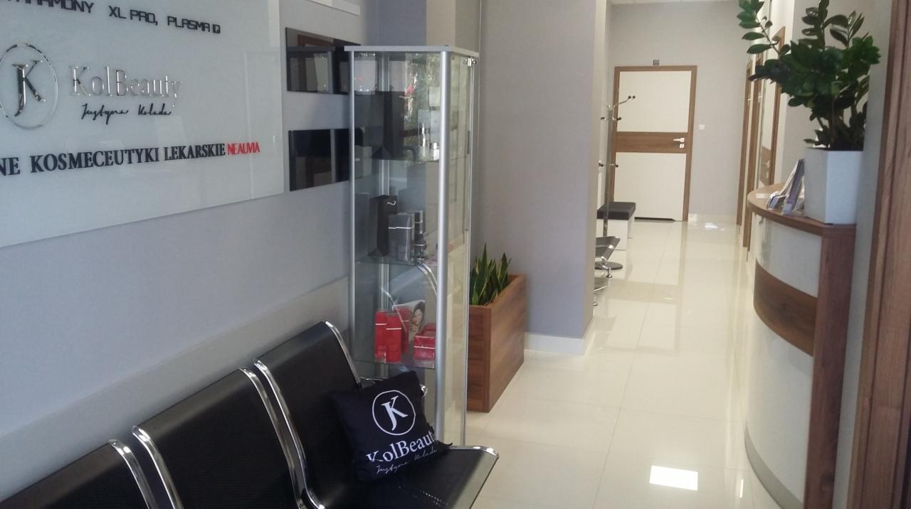 Wnętrze gabinetu: poczekalnia z czarnymi krzesłami, szklana witryna z kosmetykami, recepcja i logo firmy na ścianie.