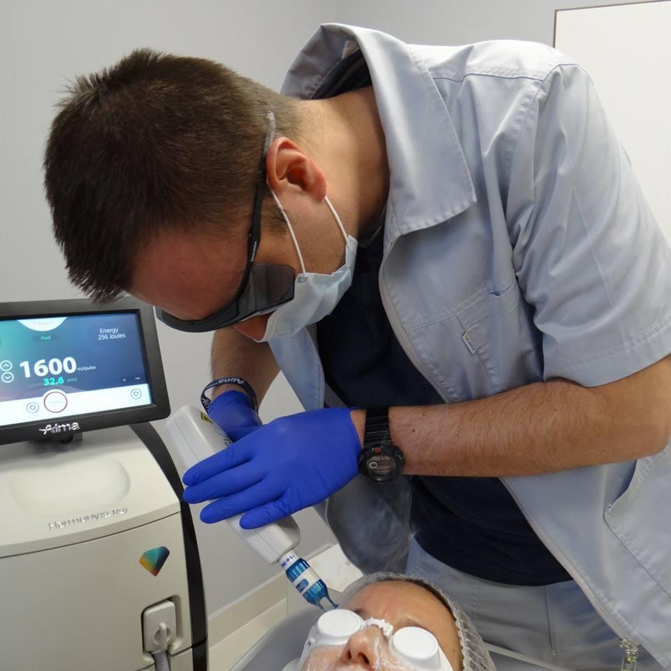Lekarz w okularach ochronnych wykonuje zabieg laserowy na twarzy pacjentki z osłoniętymi oczami, widoczny ekran urządzenia Alma z parametrami zabiegu.