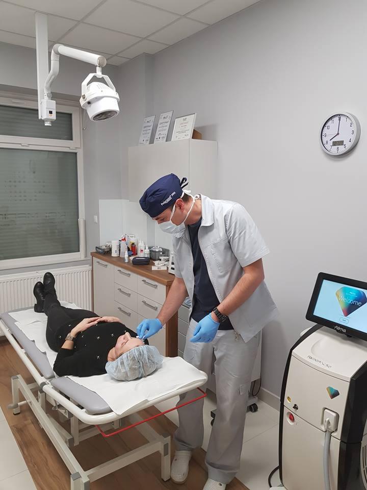 Lekarz w rękawiczkach i maseczce aplikuje preparat na twarz pacjentki leżącej na kozetce w gabinecie zabiegowym, widoczny sprzęt medyczny i zegar na ścianie.