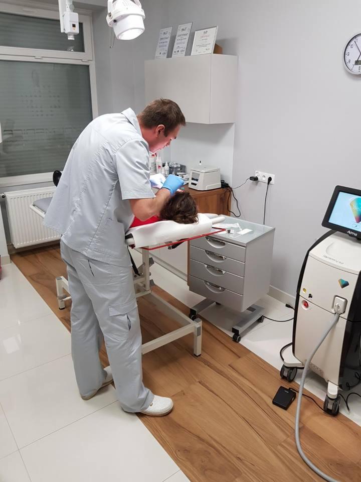 Lekarz w niebieskich rękawiczkach wykonuje zabieg na pacjentce leżącej na białym łóżku zabiegowym w gabinecie, obok widoczny laser Alma Harmony XL Pro.