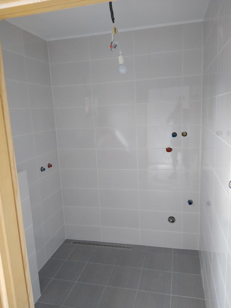 Wykończona łazienka z szarymi płytkami podłogowymi i jasnymi płytkami ściennymi, widoczne przyłącza wodne i odpływ liniowy, surowa żarówka zwisająca z sufitu.