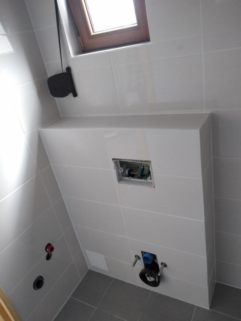 Świeżo wykafelkowana toaleta podtynkowa w łazience z szarą podłogą i oknem dachowym, widoczne przyłącza hydrauliczne.