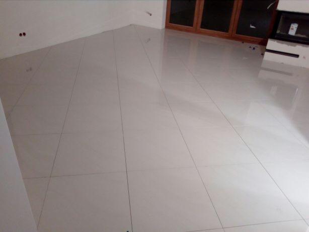 Błyszcząca, jasna podłoga wyłożona dużymi, kwadratowymi płytkami ceramicznymi z minimalną fugą, odbijająca światło od okna i kominka, w trakcie realizacji.