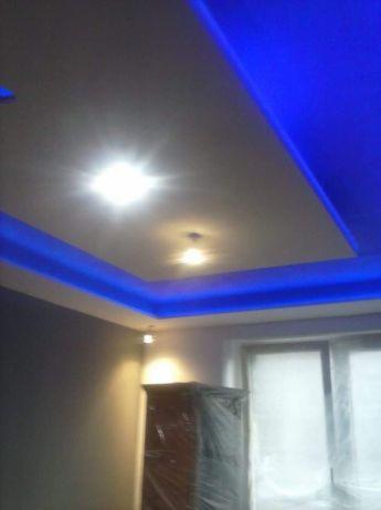 Biały sufit z niebieskim oświetleniem LED wzdłuż krawędzi, widok z dołu, z lampami punktowymi i meblem zabezpieczonym folią w tle.