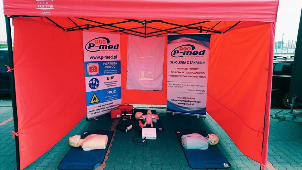 Stanowisko szkoleniowe pod czerwonym namiotem z manekinami do ćwiczeń pierwszej pomocy, torbami medycznymi i banerami informacyjnymi firmy P-med.