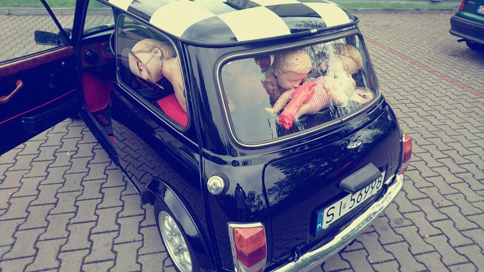 Czarny Mini Cooper z szachownicą na dachu, wypełniony manekinami i lalkami, zaparkowany na brukowanym placu. Manekin w okularach siedzi na czerwonym siedzeniu, a lalki leżą na tylnej półce.
