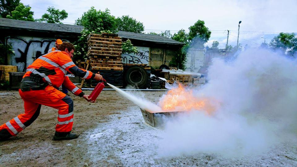 Osoba w pomarańczowym kombinezonie ochronnym gasi ogień w metalowym pojemniku za pomocą gaśnicy proszkowej podczas symulacji pożaru na placu.
