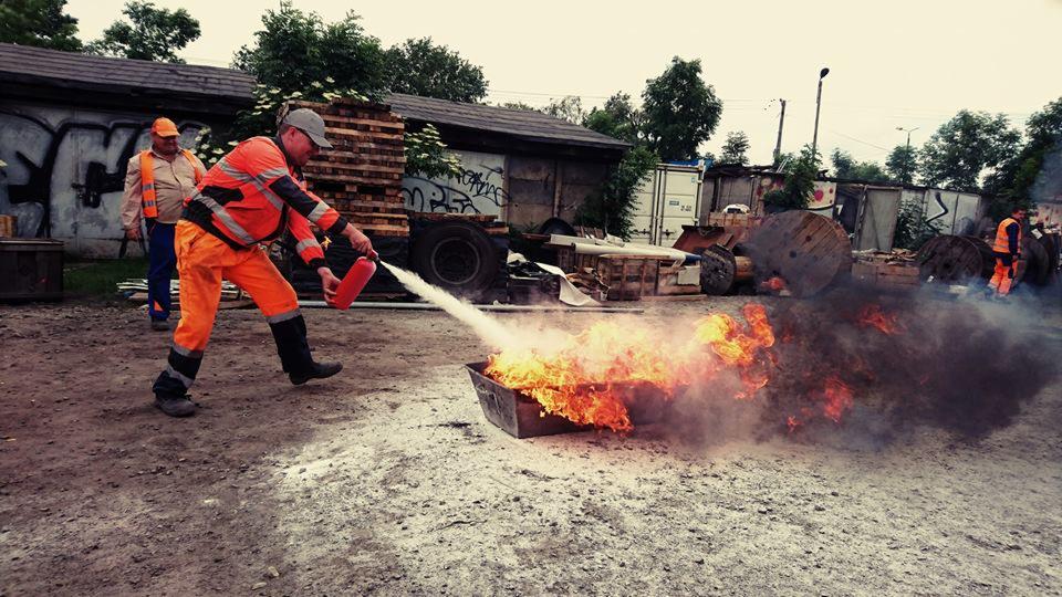 Mężczyzna w pomarańczowym kombinezonie gasi ogień w metalowej skrzyni za pomocą gaśnicy, na ziemi rozsypany biały proszek, w tle budynki i składowisko materiałów.