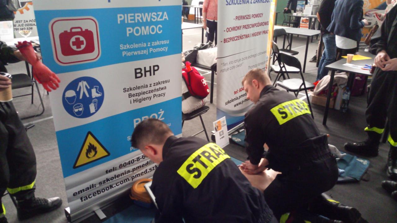 Instruktorzy straży pożarnej demonstrujący resuscytację krążeniowo-oddechową na manekinie podczas targów, obok banery informacyjne z zakresu pierwszej pomocy, BHP i ochrony przeciwpożarowej.