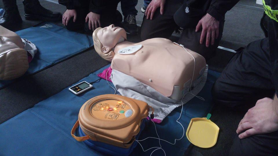 Manekin do ćwiczeń resuscytacji krążeniowo-oddechowej z podłączonym defibrylatorem AED i elektrodami na klatce piersiowej, leżący na niebieskiej macie, otoczony przez osoby w trakcie szkolenia.
