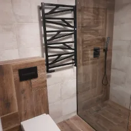 Nowoczesna łazienka z płytkami imitującymi drewno, czarnym grzejnikiem drabinkowym, kabiną prysznicową typu walk-in i podtynkową toaletą z czarnym przyciskiem.