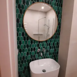 Elegancka łazienka z umywalką na postumencie, lustrem w złotej ramie i ścianą wyłożoną zielono-czarnymi płytkami w kształcie rybiej łuski.