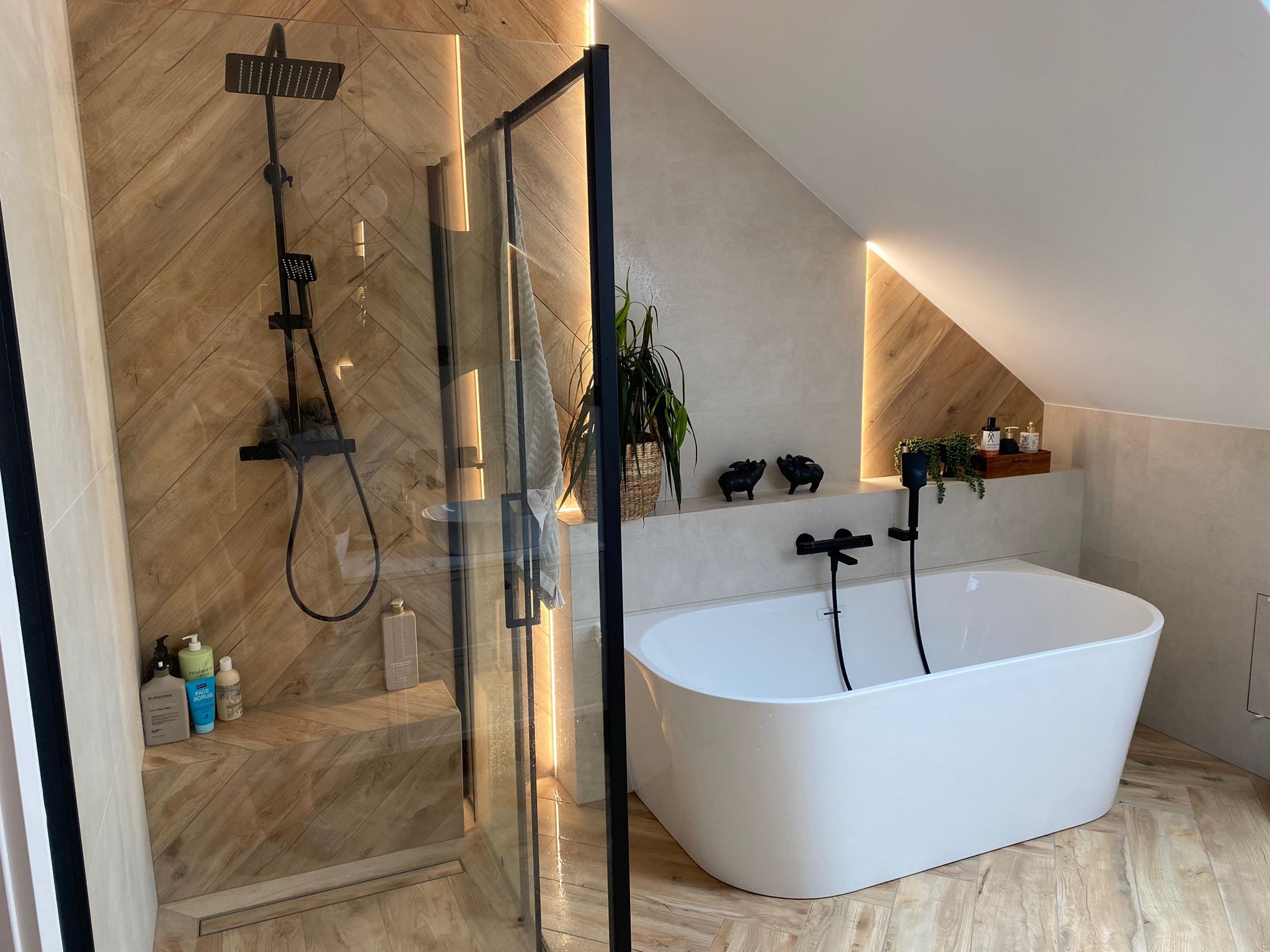 Nowoczesna łazienka na poddaszu z drewnopodobnymi płytkami ułożonymi w jodełkę, czarną armaturą i oświetleniem LED. Kabina prysznicowa typu walk-in i wolnostojąca wanna.