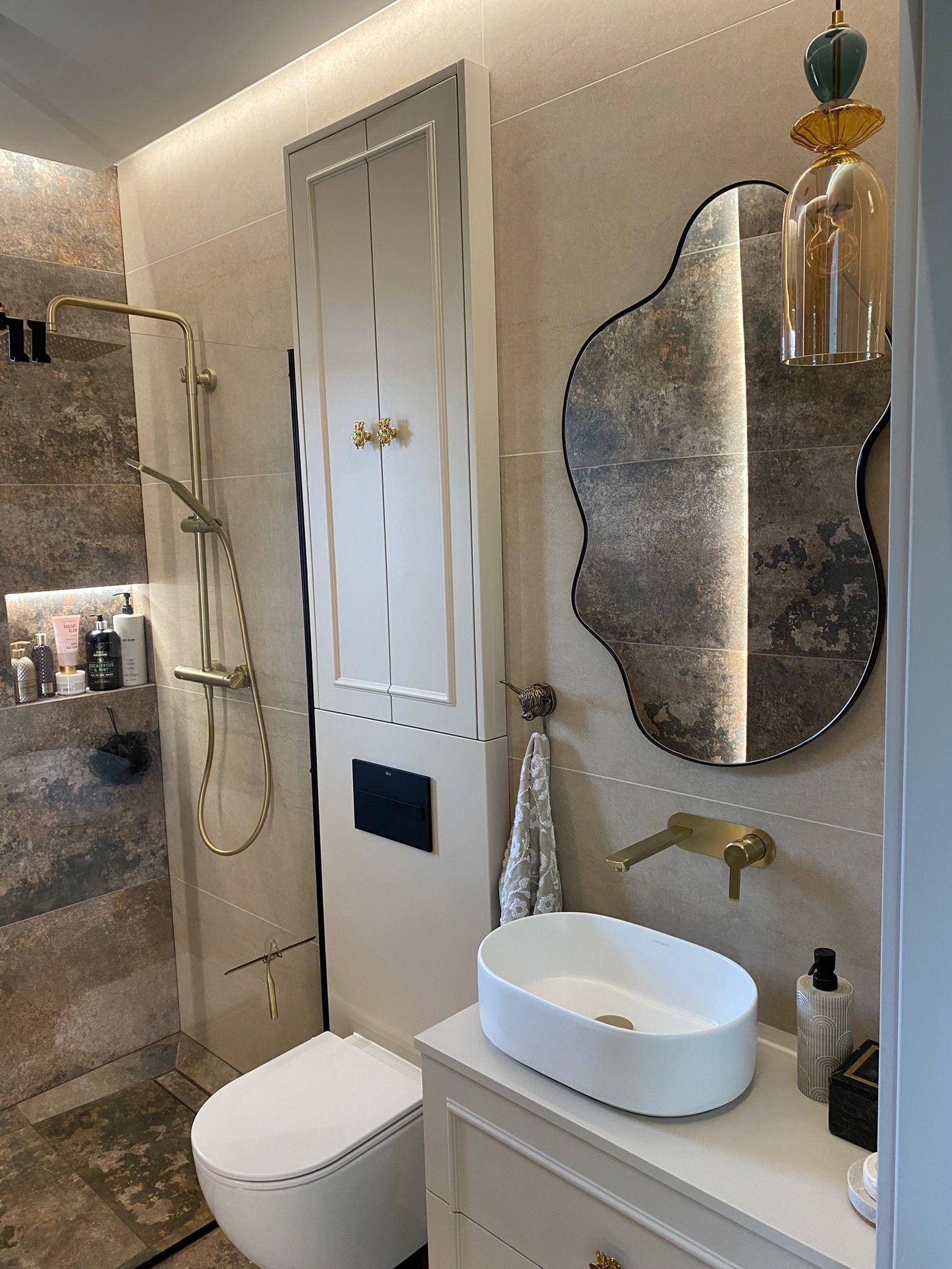 Elegancka łazienka z kabiną prysznicową typu walk-in, umywalką nablatową i lustrem o nieregularnym kształcie. Złote akcenty armatury i oświetlenia.