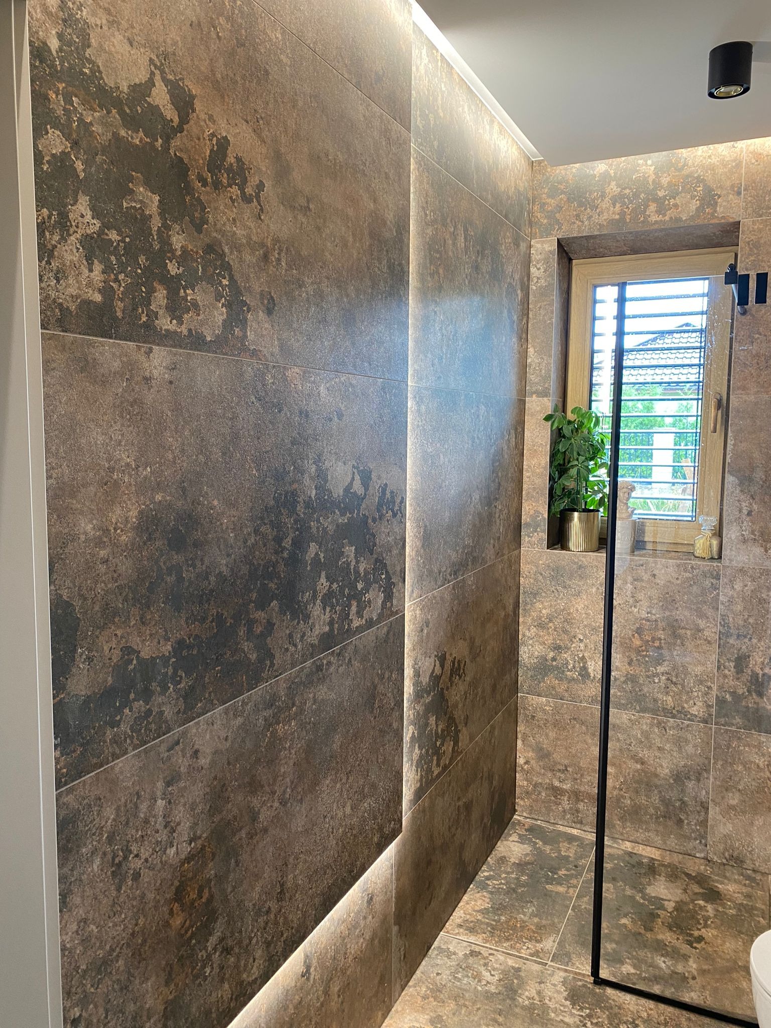 Nowoczesna łazienka z dużymi, brązowymi płytkami imitującymi kamień, podświetleniem LED i oknem z roletami. Minimalistyczny design z rośliną w złotej doniczce.